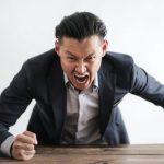 Bullying - Expressive angry businessman in formal suit looking at camera and screaming with madness while hitting desk with fist