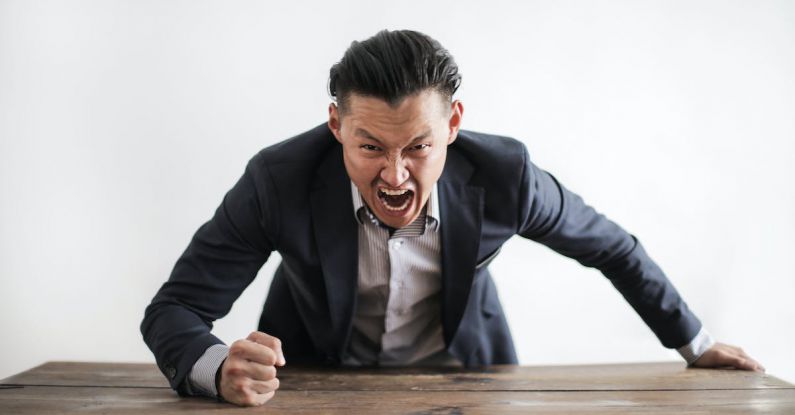 Bullying - Expressive angry businessman in formal suit looking at camera and screaming with madness while hitting desk with fist
