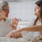 Home Care - Calm senior woman and teenage girl in casual clothes looking at each other and talking while eating cookies and cooking pastry in contemporary kitchen at home