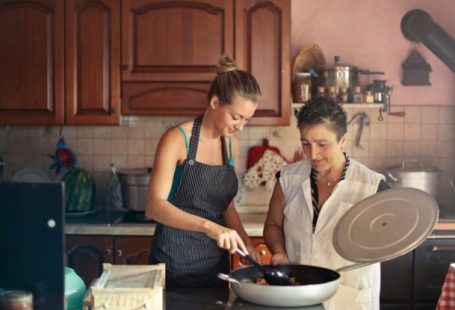 Senior Nutrition - Daughter and senior mother standing at table in kitchen and stirring dish in frying pan while preparing food for dinner