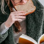 Smart Snacking - Concentrated female wearing warm coat and eyeglasses eating tasty bread while reading interesting book on white background in light room