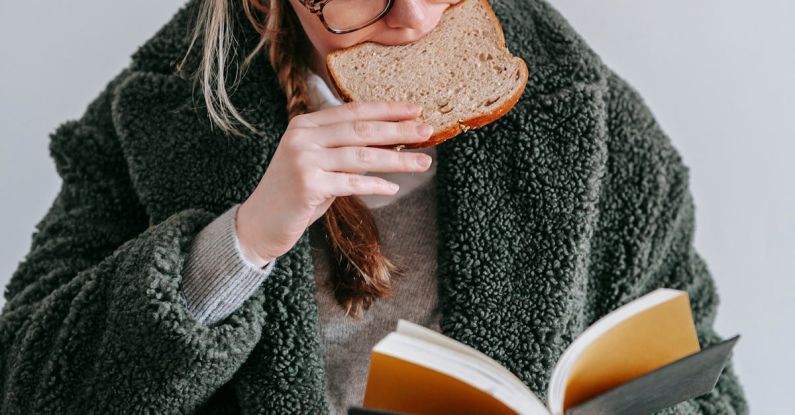 Smart Snacking - Concentrated female wearing warm coat and eyeglasses eating tasty bread while reading interesting book on white background in light room