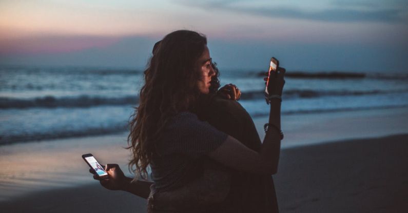 Distant Relatives - Couple hugging and using smartphone near sea on sunset